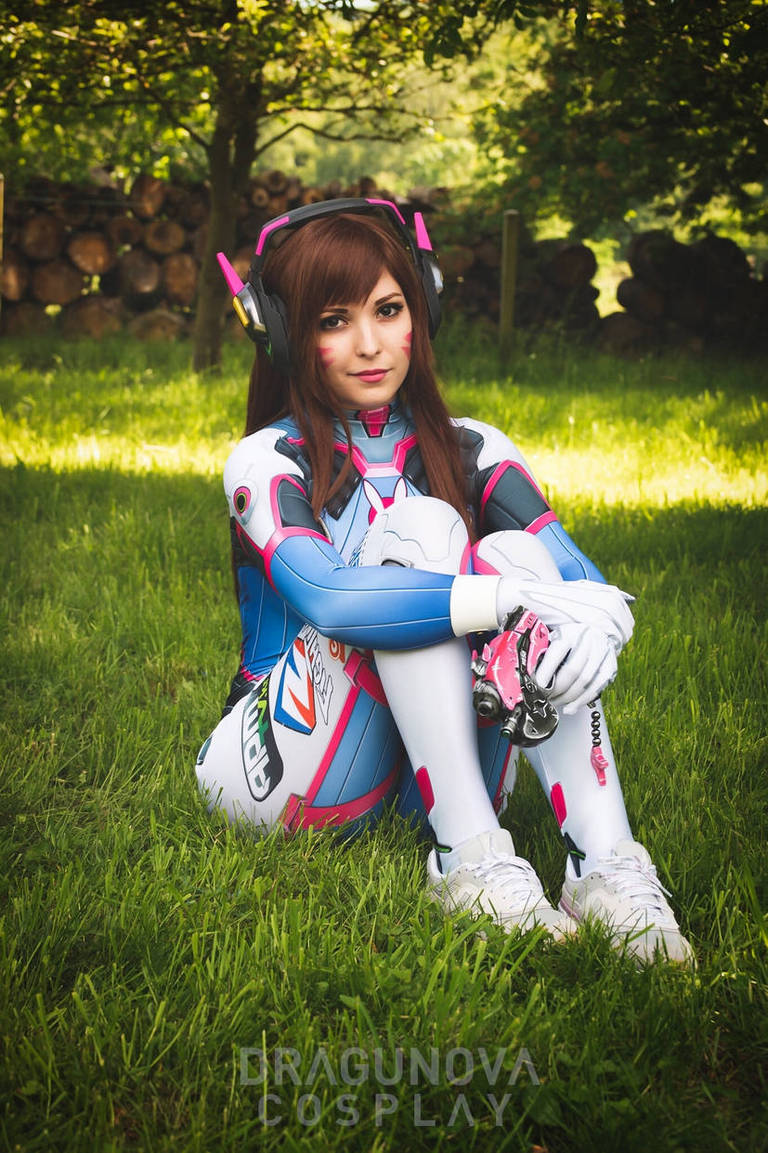 A person dressed in a colorful cosplay outfit inspired by a video game character, sitting on green grass. The costume features a bodysuit with shades of blue, pink, and white, along with large headphones.