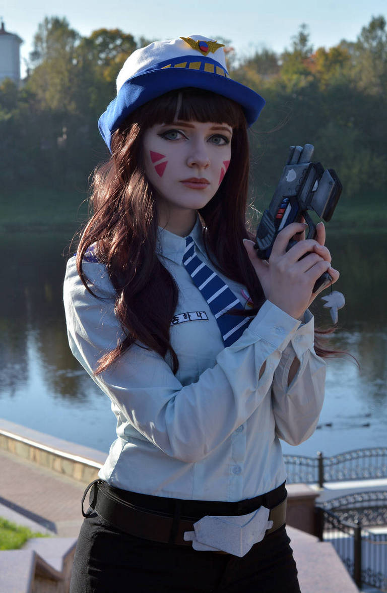 A woman in a blue cap and shirt holding a toy gun, with playful makeup resembling a character, posing by a riverbank in a park setting.