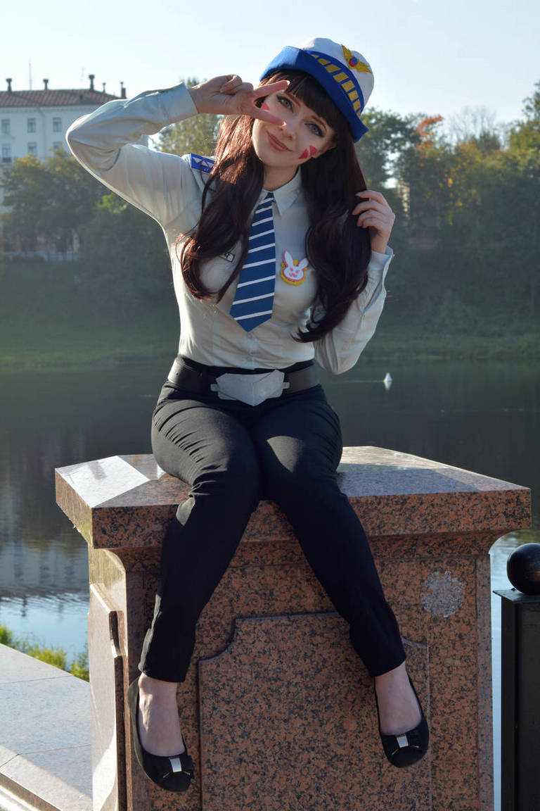 A person dressed in a blue and gray police-themed costume with a hat, sitting on a stone pedestal and making a peace sign outdoors, with a body of water and trees in the background.