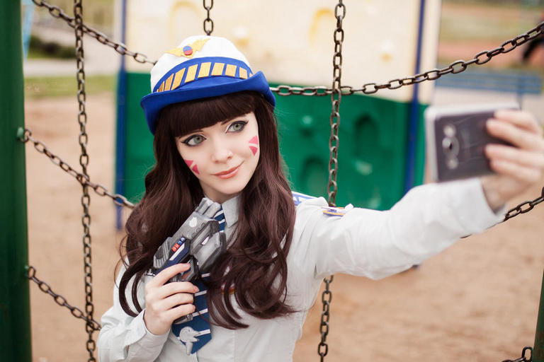 A woman dressed as a video game character, wearing a hat and holding a toy gun, takes a selfie in a playground.