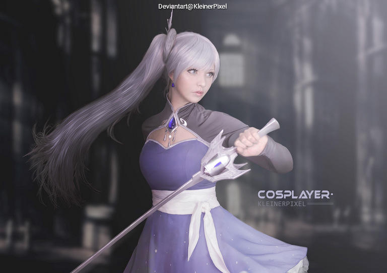 A cosplayer dressed in a light purple gown with a silver robe, holding a magical staff with a crescent-shaped top. The character has long silver hair styled in a ponytail and is posing in a dramatic environment.