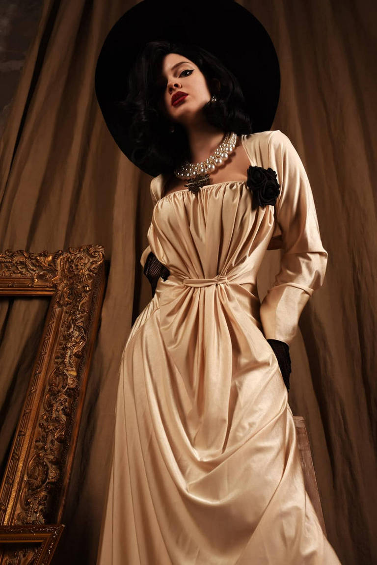 A woman in a vintage-inspired, elegant satin gown poses dramatically, wearing a large black hat and a pearl necklace, against a backdrop of draped fabric and ornate picture frame.