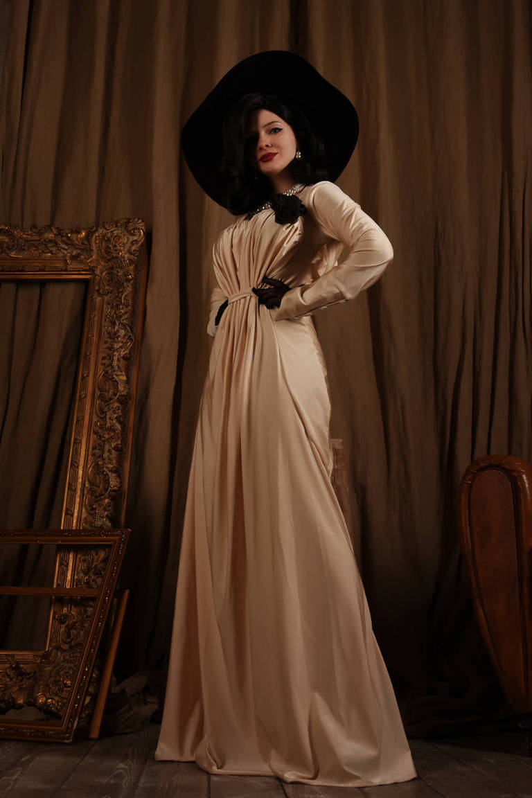 A woman poses elegantly in a long, flowing beige dress with a black flower detail, complemented by a large black hat. The background features rich brown drapery and ornate picture frames, creating a classic and sophisticated atmosphere.