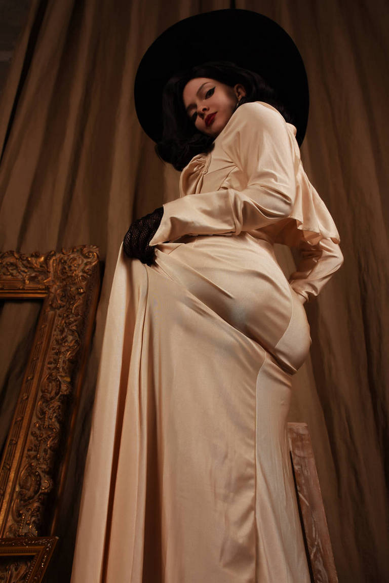 A model poses dramatically in a silky, cream-colored dress, wearing a large black hat and black gloves, with an ornate frame in the background.