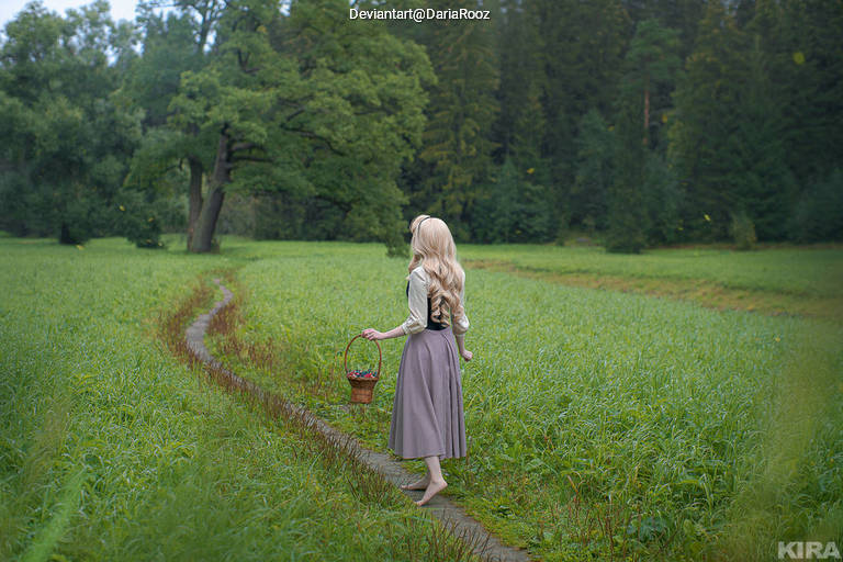 A woman with long blonde hair walks barefoot along a dirt path in a green field, holding a basket. She is dressed in a light blouse and a long skirt, with trees in the background under a cloudy sky.