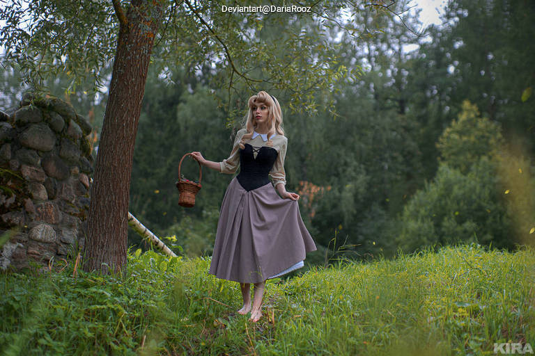 A young woman in a medieval-style dress stands barefoot on a grassy area, holding a small basket and looking thoughtfully into the distance. Surrounding her are trees and a stone wall, creating a serene outdoor setting.
