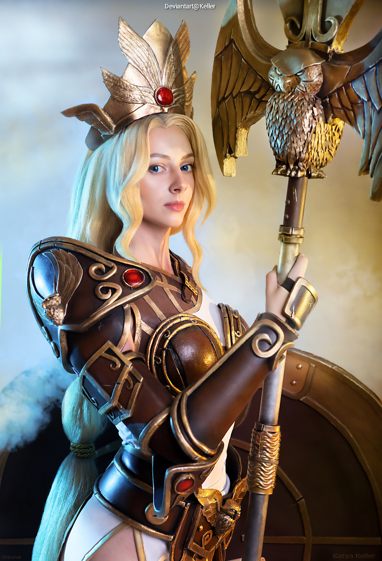 A cosplayer dressed as a fantasy warrior, featuring elaborate armor with golden accents and red gemstones, holding a large ornate staff topped with an eagle. The character has long blonde hair and is posed against a vibrant background.