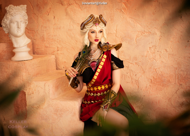 A woman dressed in vibrant cosplay attire inspired by ancient themes, featuring a red and green outfit with intricate armor and a decorative headdress. She holds a peacock feather and poses in front of a textured orange wall with a classical bust in the background.