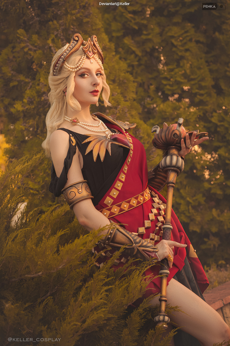 A woman dressed in an elaborate, regal costume with gold and red details, holding a staff, posing elegantly in front of greenery.