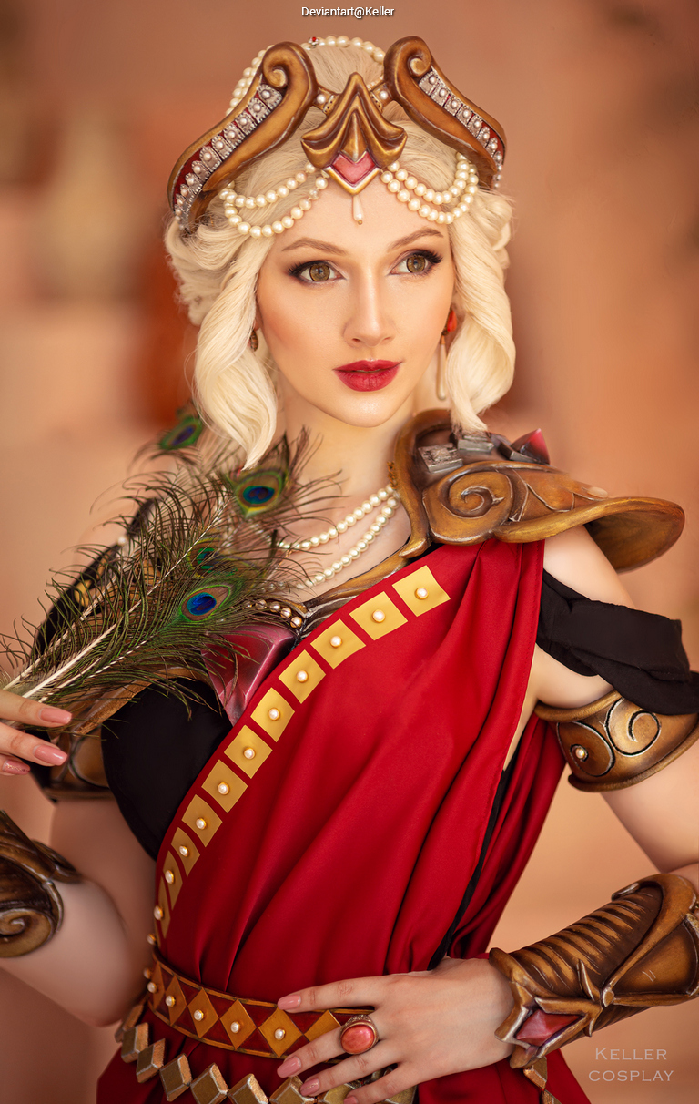 A woman in a red toga-style costume with intricate golden armor and a peacock feather, posing elegantly against a soft background.