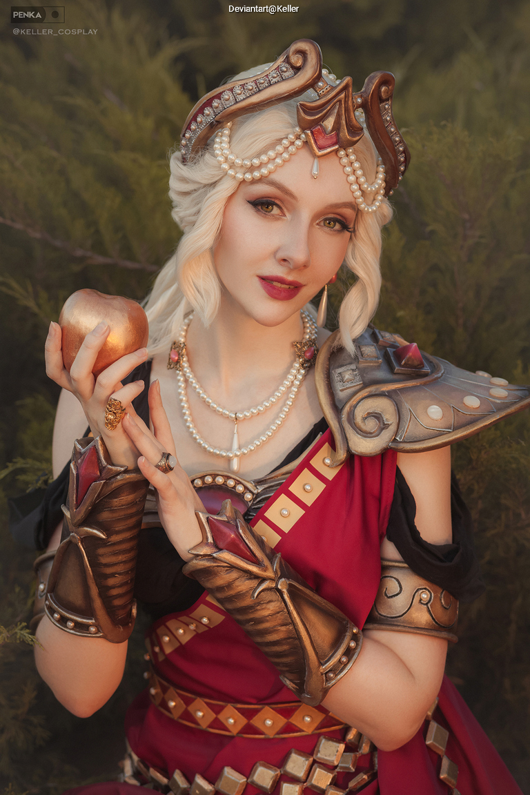 Woman dressed in a red and gold costume resembling a historical or fantasy character, holding an apple and adorned with elaborate jewelry and armor.