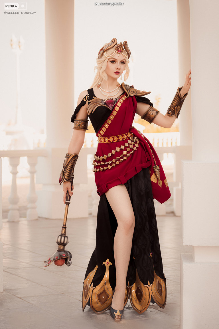 A person in a sophisticated warrior costume featuring a red and black dress, adorned with gold accents and armor. They hold a staff with a decorative top, and the setting appears to be an elegant architectural space.