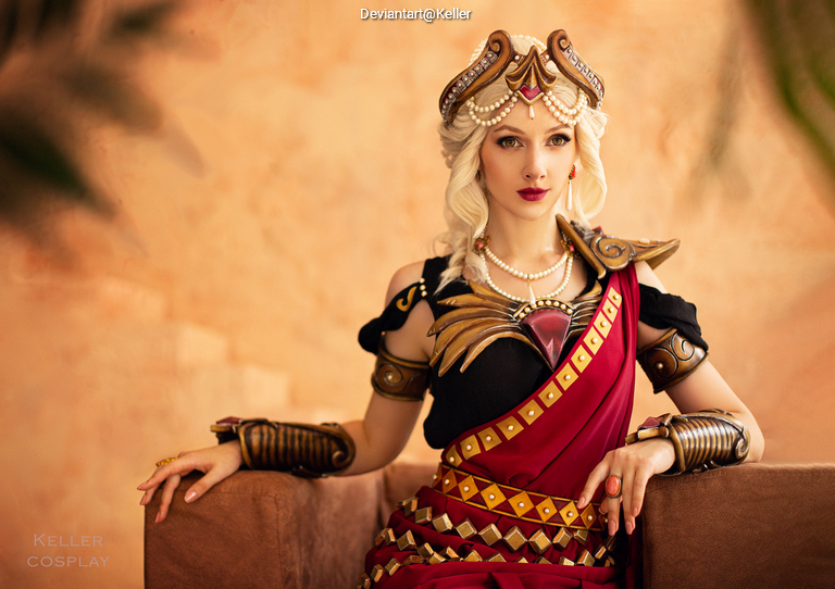 A cosplayer dressed as a regal character, featuring a detailed costume with armor, a flowing red and black robe, and intricate accessories, seated on a brown couch against a warm-colored backdrop.