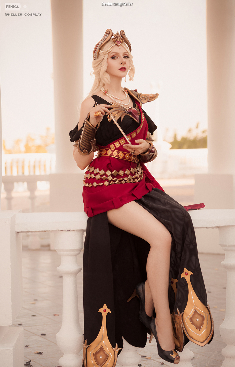A person dressed in intricate cosplay attire, featuring ornate armor, a flowing skirt, and decorative accessories, posing confidently while sitting on a ledge in a well-lit environment.