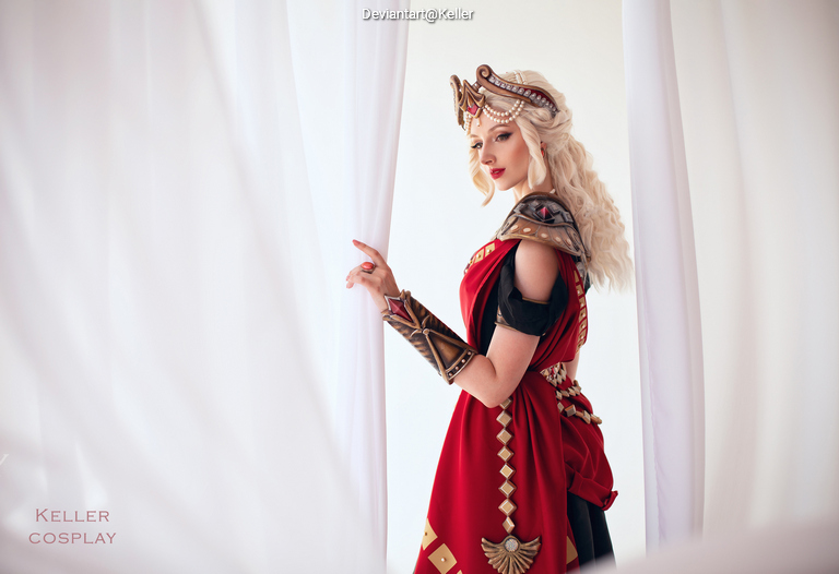 A woman in an elaborate costume stands behind sheer curtains, showcasing intricate armor and a flowing red garment.