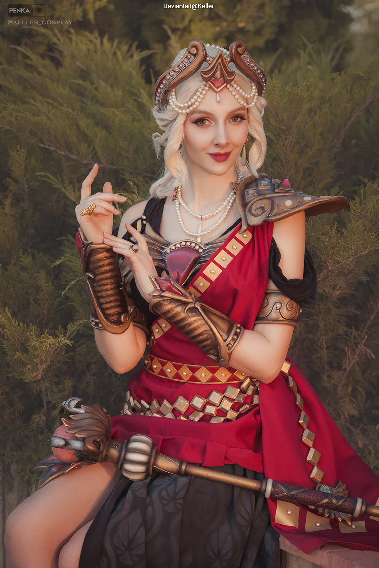 A person dressed in elaborate cosplay attire featuring a red and black outfit with armor details and accessories, sitting amidst greenery and posing elegantly.