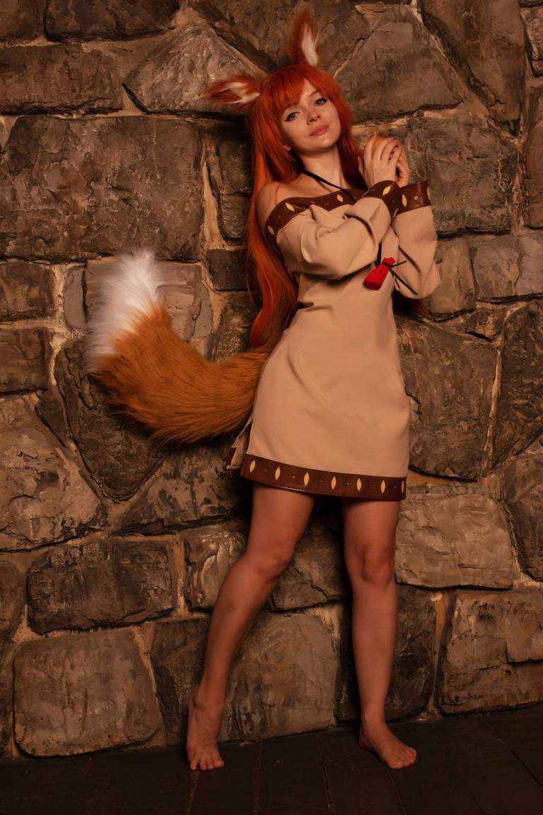 A person dressed in a fox costume with a long tail, standing in front of a stone wall. They have long orange-red hair and are posing with their hands clasped together.