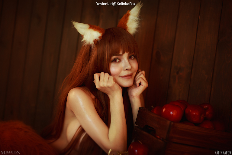 A woman with long brown hair and fox ears poses playfully in a cozy setting, surrounded by red apples and wooden decor.