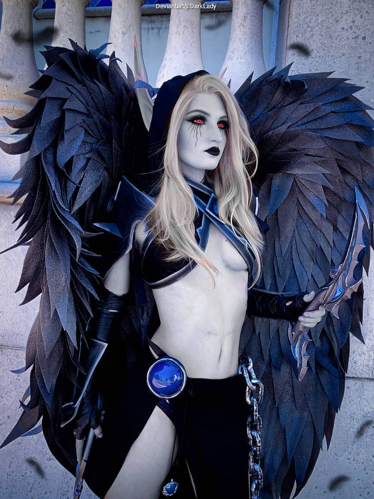 A woman in an elaborate dark costume with large black feathered wings, long blonde hair, and striking red eyes. She holds a weapon in one hand and has a mystical emblem on her outfit, standing against a stone backdrop.
