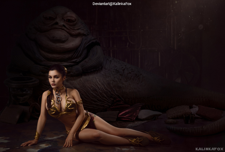 A woman in a gold costume poses in front of a large, character resembling Jabba the Hutt, in a dimly lit environment.