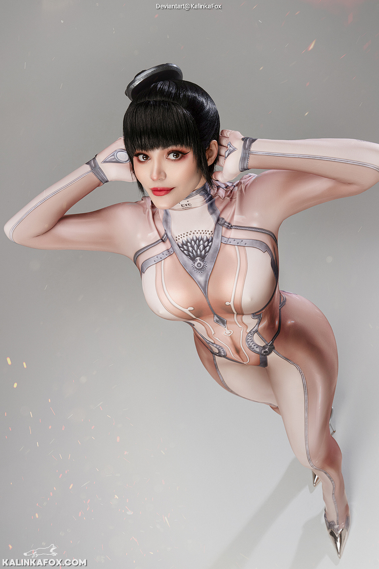 A woman in a futuristic, skin-tight outfit poses with her arms behind her head, showcasing a confident expression against a soft gray background.