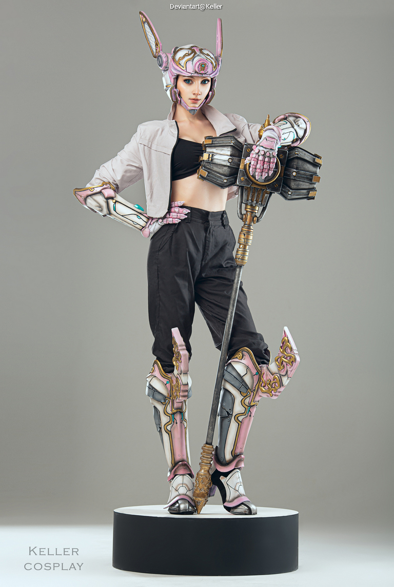 A cosplayer in a detailed, sci-fi inspired costume with pink and silver armor, holding a large weapon, standing on a circular podium.