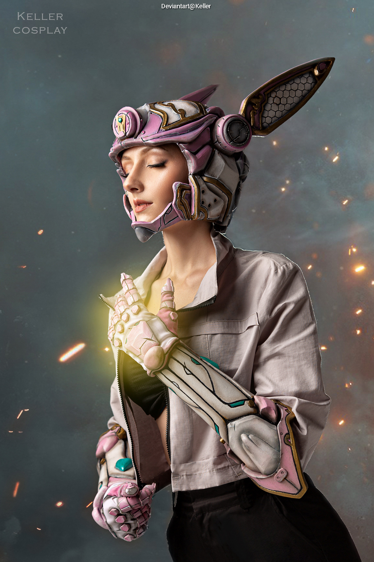 A person cosplaying as a character with a bunny-themed helmet and robotic arm, wearing a grey jacket and striking a pose against a softly lit background with sparks.