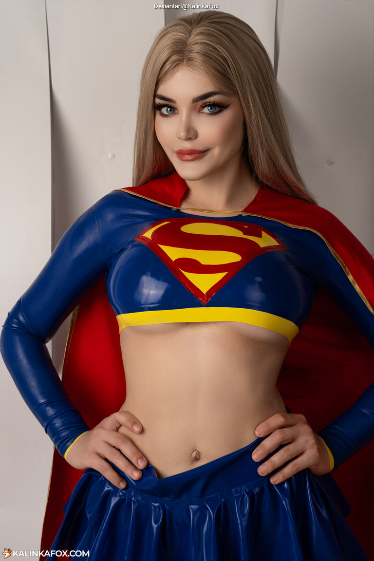 A woman in a superhero costume, featuring a blue top with a yellow logo and a red cape, posing confidently.