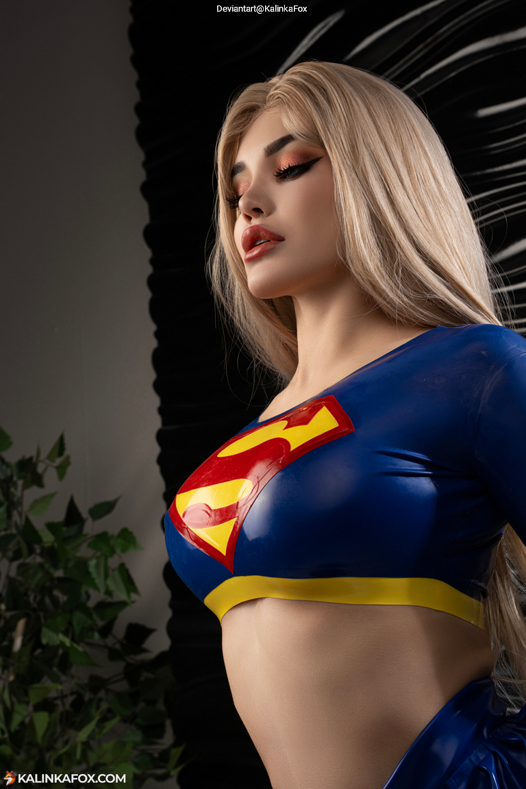 A person dressed as a superhero in a blue and yellow costume featuring a large emblem on the chest, posing confidently with a side profile against a dark background.