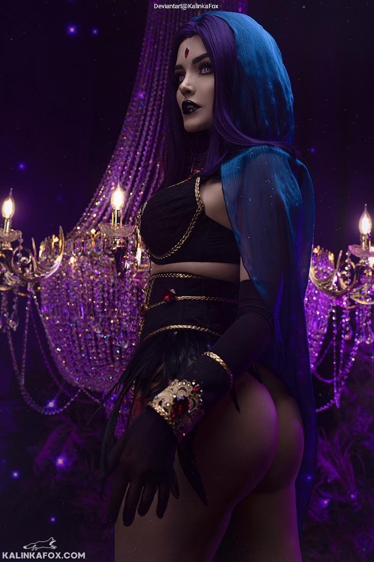 A woman with long purple hair and dramatic makeup poses under a chandelier, wearing a black outfit with a blue hooded cape, against a dark purple background.
