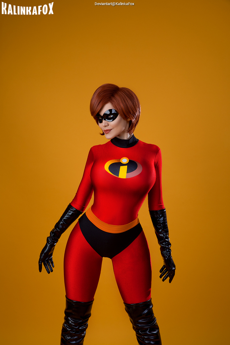 A person dressed as a superhero in a red and black costume, featuring an emblem with a yellow circle, posing against a bright orange background.