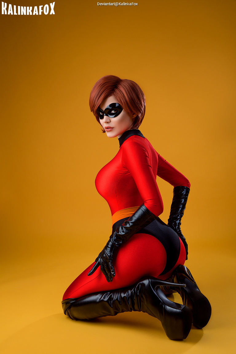 A woman dressed in a superhero costume with a red bodysuit, black gloves, and a black mask, posing on her knees against a yellow background.