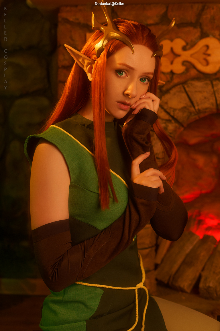 A person dressed in fantasy-themed cosplay, featuring long red hair and pointed ears, poses thoughtfully with a hand on their face. The character is wearing a green outfit with dark sleeves, set against a softly lit, rustic background with a fireplace.