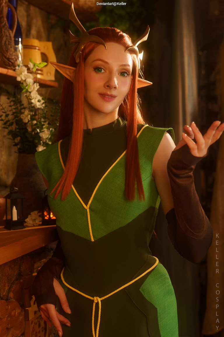 A person cosplaying as an elven character with long reddish-brown hair and pointed ears, wearing a green outfit with yellow accents, standing in a cozy environment.