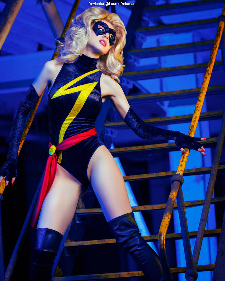 A person dressed as a superhero in a black and yellow costume with a lightning bolt design, posing dramatically on a staircase with blue lighting.