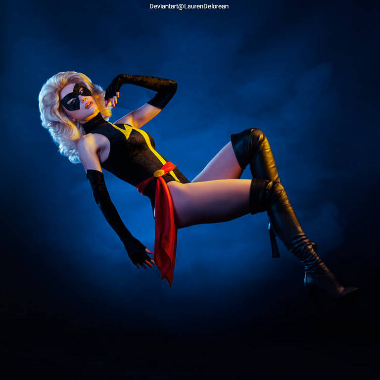 A superhero character in a black costume with a red sash and high black boots, posed dramatically against a blue smoky background.