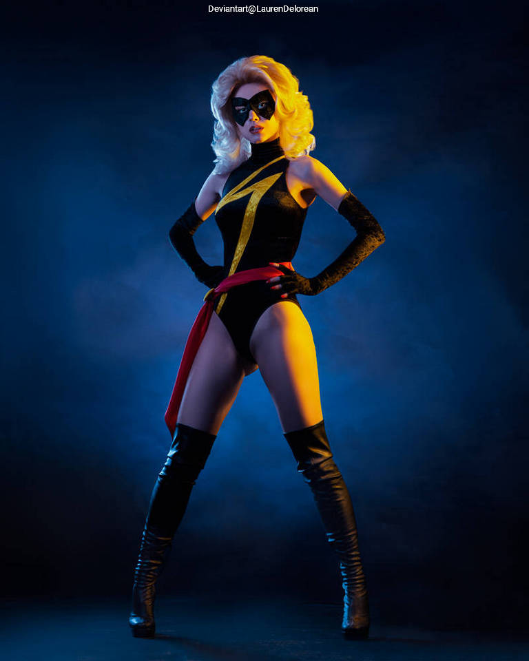 A superheroine in a black costume with yellow accents and a red sash, posing confidently against a dramatic blue backdrop.