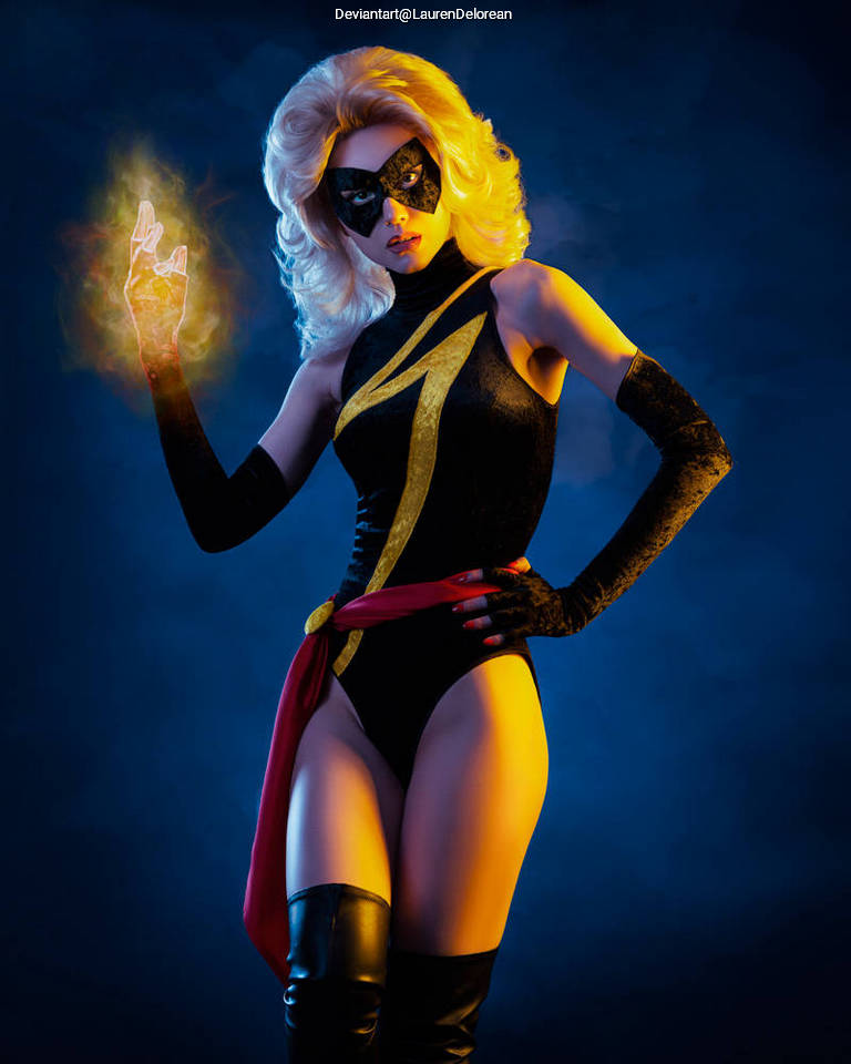 A person in a superhero costume with blonde hair and a mask, holding a glowing orange energy effect in one hand against a blue background.