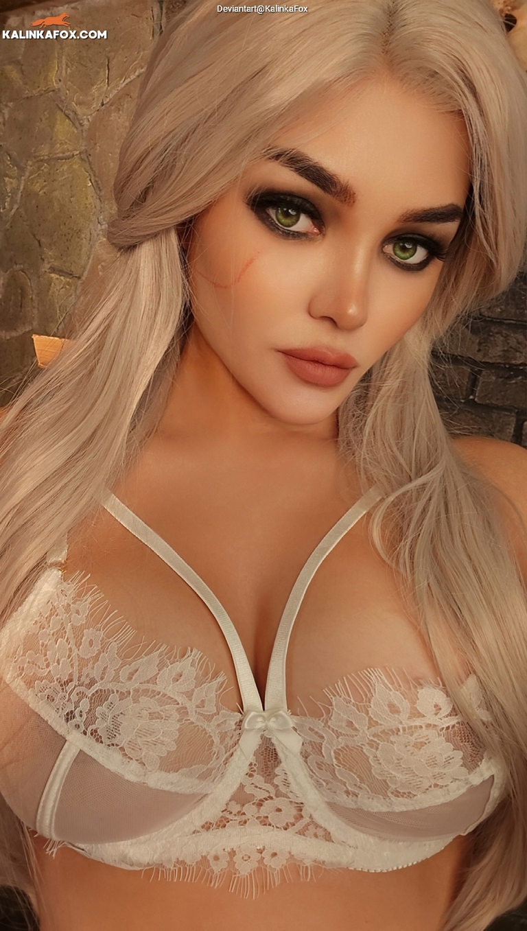 Close-up portrait of a woman with long, platinum blonde hair and striking green eyes, wearing a white lace bra. The background features a textured stone wall.