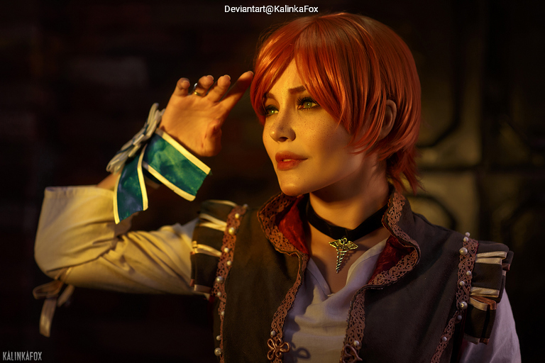 A person with short orange hair and green eyes wearing a costume with a gray vest and a blue ribbon, posing with one hand raised near their face.