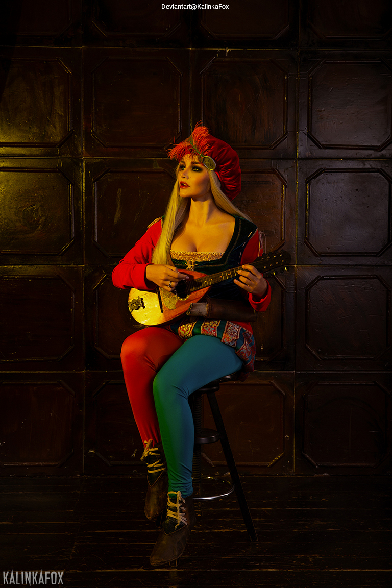 A woman dressed in colorful attire with a red hat sits on a stool, holding a small string instrument, set against a dark wooden backdrop.
