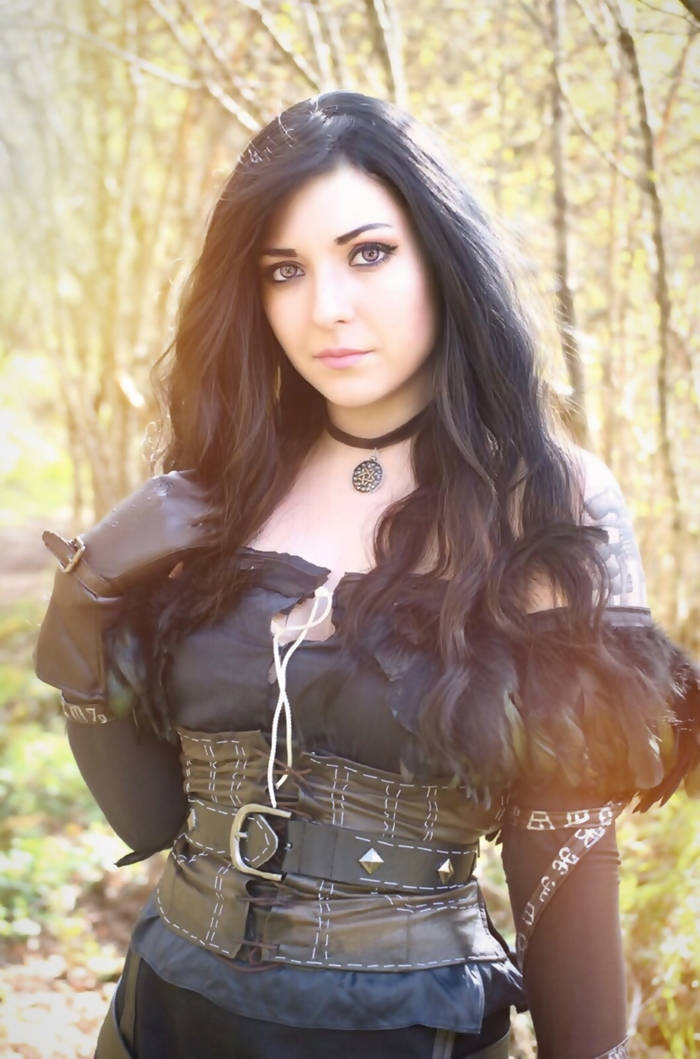 A woman with long black hair and striking blue eyes stands in a sunlit forest. She wears a dark, fashionable outfit with a corset-style top and a choker necklace.