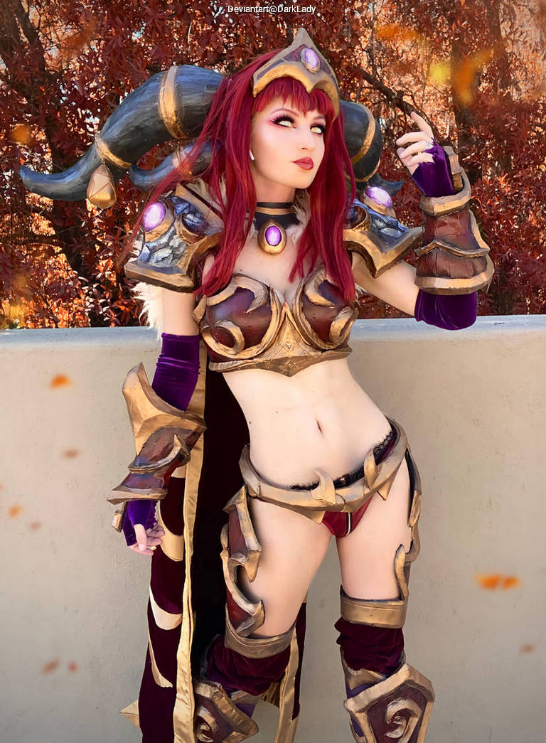 A person in elaborate fantasy-themed armor with horns and vibrant red hair posing in front of a backdrop of autumn leaves.