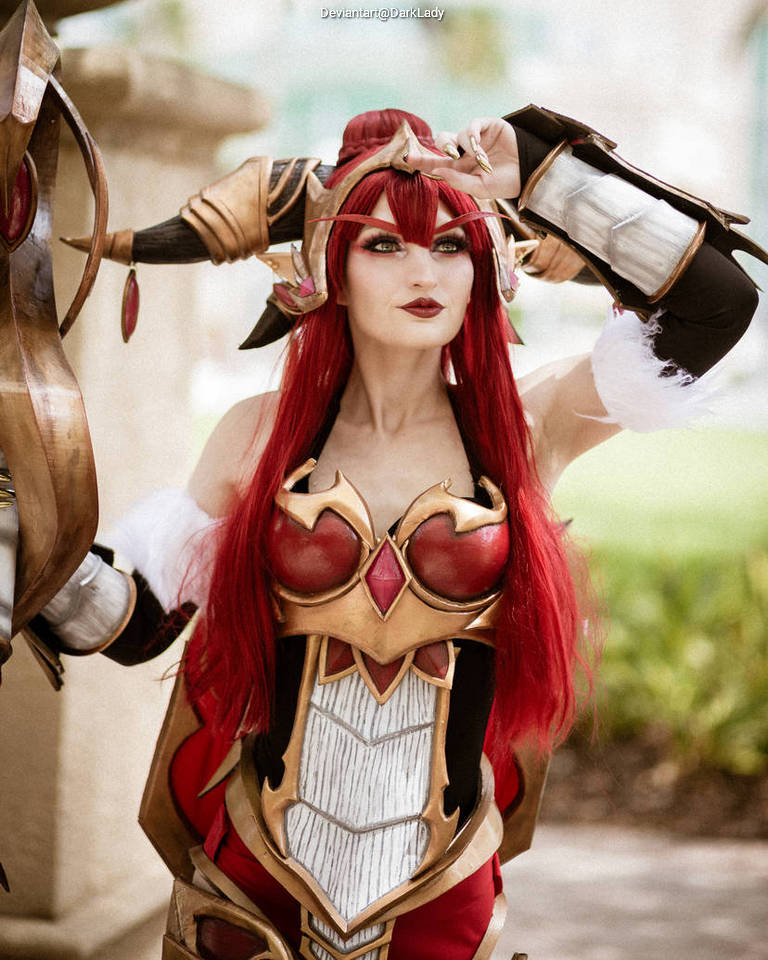 A cosplayer dressed in elaborate armor with red accents and long red hair, striking a playful pose outdoors.