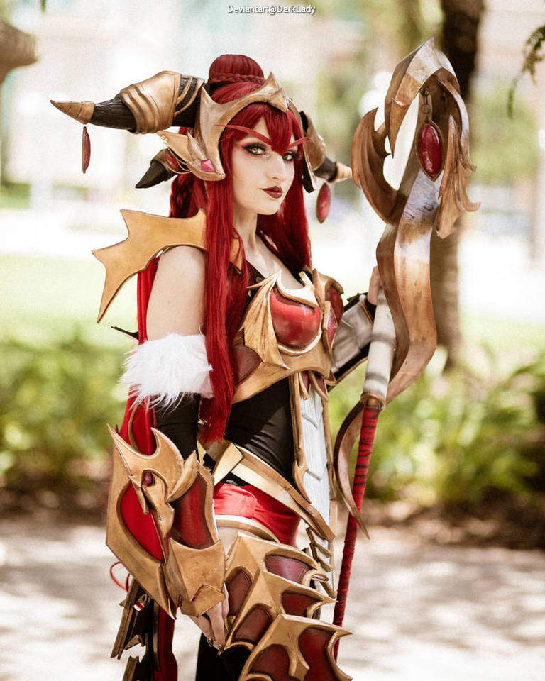 A cosplayer dressed in an elaborate fantasy costume with red hair, striking armor, and a staff, posing outdoors.