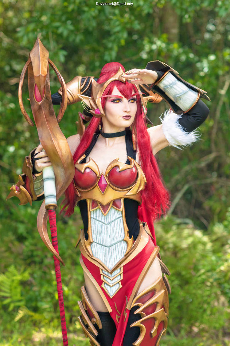 A person in an elaborate costume featuring armor-like details and vibrant red hair, striking a pose outdoors with greenery in the background.