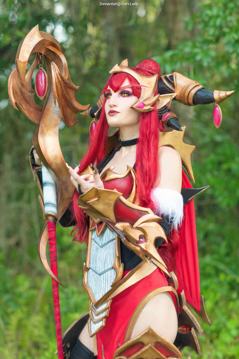 A female cosplayer dressed in elaborate red and gold armor, holding a staff and posing against a green natural background.