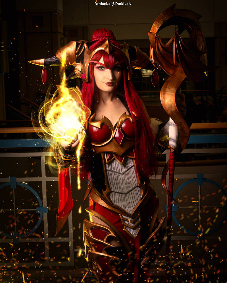 A female cosplayer in an elaborate fantasy armor costume, holding a glowing orb of fire, surrounded by sparks and colorful lighting.