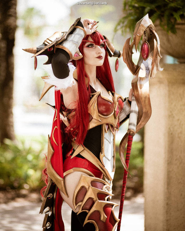 A female cosplayer in an elaborate costume featuring red and gold armor, holding a staff with decorative elements, set against an outdoor background.