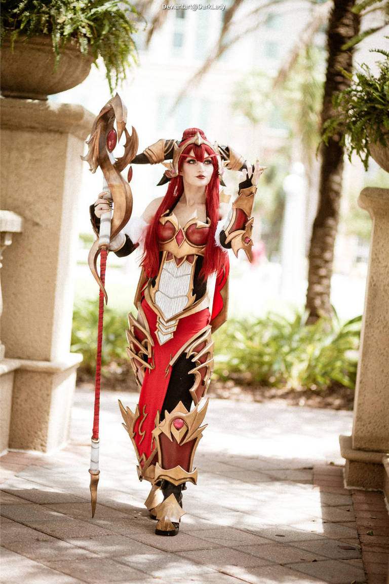 A cosplayer dressed in a red and gold fantasy costume, holding a staff. She has long red hair and is posing outdoors among greenery.